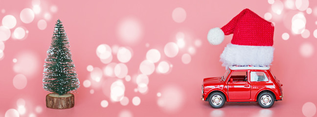 Red toy car with a santa hat, next christmas tree on pink paper background. Winter delivery, xmas, happy new year 2020 celebration concept. greeting card, mockup, copy space, place for text, template