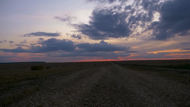 Timelapse of cloudy orange sunset sky over harvest field with haystack