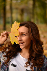 Happy young attractive woman with curly hair, covering her face with yellow autumn maple leaf in the park. Cute girl enjoying warm weather. Autumn mood. Enjoy season