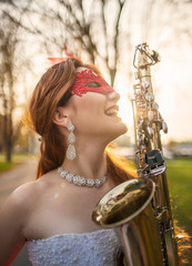 A smiling girl in a wedding dress with a red lace mask, in sun rays, on background of autumn park,...