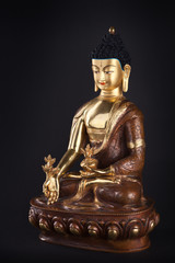 The gilded statue of Buddha Medicine in traditional clothes sitting in a lotus pose with flower of a medical plant of an arur. Isolated on black background.