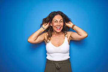 Obraz na płótnie Canvas Middle age senior woman with curly hair standing over blue isolated background Smiling pulling ears with fingers, funny gesture. Audition problem