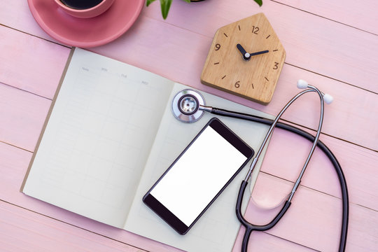 Mockup image of medical stethoscope, mobile smart phone with blank white screen,empty weekly diary notebook,wooden clock and pink cup of coffee isolated on sweet color wood table background.Top view  