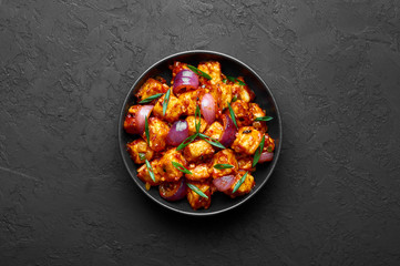 Schezwan Paneer in black bowl at dark slate background. Schezwan Paneer is indo-chinese cuisine dish with deep fried Paneer cheese, onion and Schezwan Sauce. Copy Space. Top view