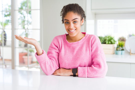Beautiful african american woman with afro hair wearing casual pink sweater smiling cheerful presenting and pointing with palm of hand looking at the camera.
