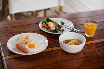 Breakfast includes bread, fried egg, various vegetable salads, sausages, fried rice, and orange juice on a brown wooden table