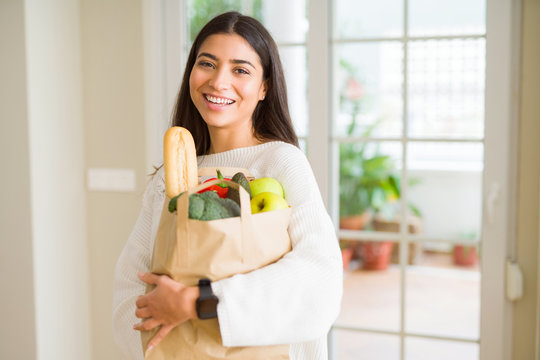 Beautiful young woman smiling holding a paper bag full of fresh groceries at home