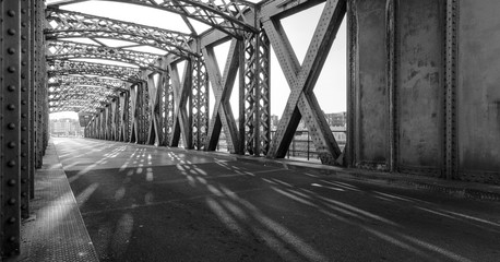 Black and white asphalt road under the steel construction of a bridge in the city on a sunny day. Evening urban scene with the sunbeam in the tunnel. City life, transport and traffic concept.	