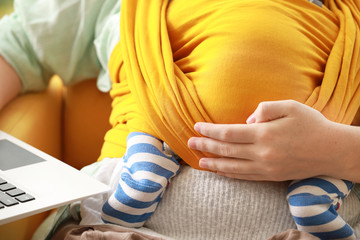 Young mother with little baby in sling using laptop at home, closeup