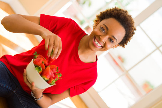 Beautiful young african woman with afro hair eating fresh strawberries sitting on the floor