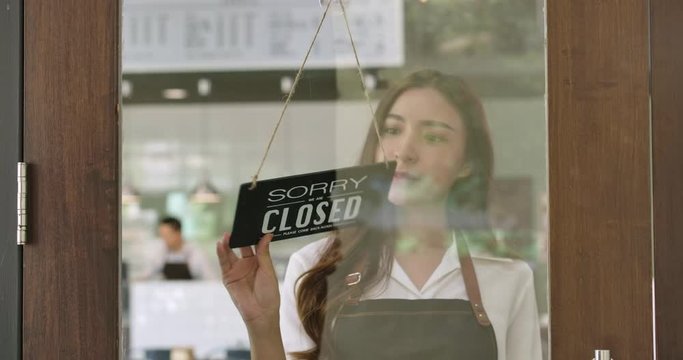 Close-up of a Attractive young asian woman turning over a "Open" sign in the morning at her cafe window and smiling looking outside waiting for clients.