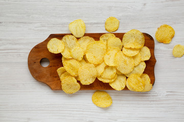 Salted wavy potato chips on a rustic wooden board on a white wooden surface, top view. Overhead, flat lay, from above.