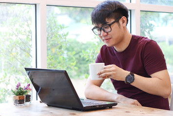 Young Asian man wearing eye glasses sitting by the window  holding cup of coffee reading mail on computer laptop