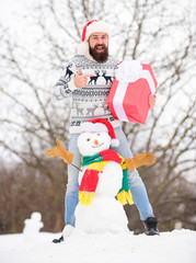 Surprise concept. Winter games. Winter activity. Winter vacation. Man made snowman. Man Santa hat having fun outdoors. Guy happy face snowy nature background. Hipster with beard hold gift box