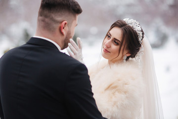 Elegant bride in white fur coat stand with handsome bearded groom in front of snow covered trees