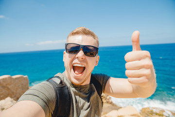 Man freelancer takes selfie photo on background of azure sea, blue sky, sunglasses and backpack. Lifestyle concept