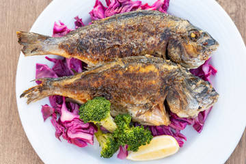 Two grilled sea fish served on a plate with red cabbage, green broccoli and lemon. Close up