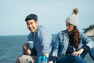 Happy Asian couple holding coffee cups on mountain peak together.