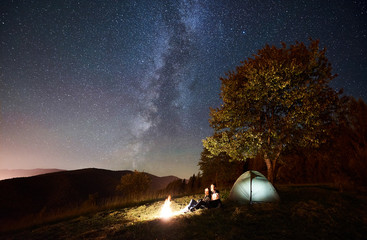 Fototapeta na wymiar Romantic couple travellers having a rest at campfire beside camp and tourist tent under amazing night sky full of stars and Milky way. On background beautiful starry sky, mountains and big tree