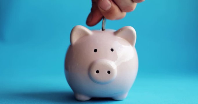 Inserting coin inside money box. Saving money using a pink coin box. Man putting coins in piggy bank of pig shape on blue background, hand closeup. Pig in center of frame.