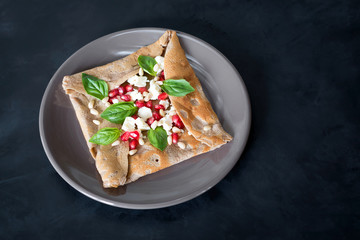 Breton galette with greek cheese, pomegranate fruit and basil on a black background.