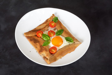 Galette bretonne with egg, tomatoes and basil on a black background.