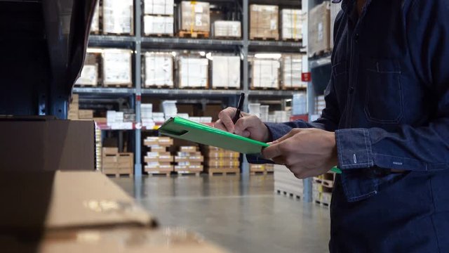 Asian manager man doing stocktaking of products management in cardboard box on shelves in warehouse using clipboard file and pen. Male professional assistant checking stock in factory.