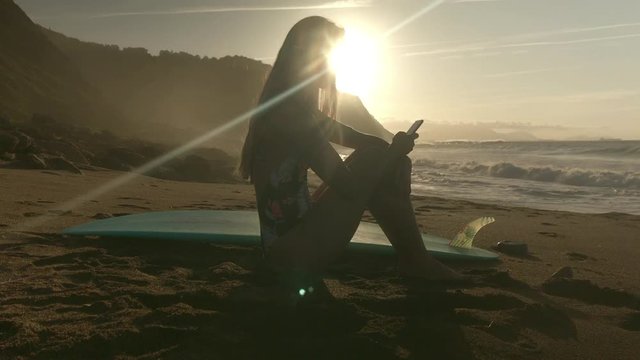 Happy young blonde surfer woman wearing a colorful swimsuit sitting in the beach with a blue surfboard using the smartphone with sunset in background