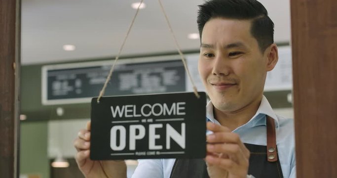 Close-up of a Attractive asian male barista turning over a "Open" sign in the morning at her cafe window and smiling looking outside waiting for clients.