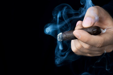 A hand holds a smoldering cigar in smoke against a dark background. Left space for text.