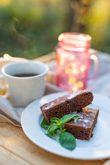 Coffee cup with a brownie cake decorated with mint on a table outdoors. Beautiful garland lights in the Mason jar on the background