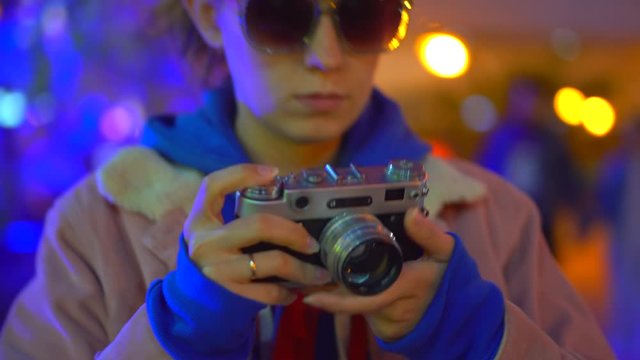 A girl in a hood and glasses stands in the neon light of the evening city lights taking pictures with a vintage camera