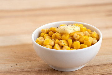 Canned sweet corn with a knob of butter and ground black pepper in a white ceramic bowl isolated on light wood. Space for text.