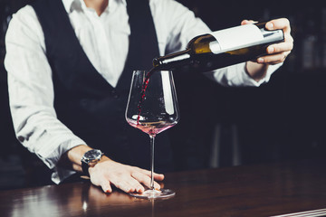 Close-up photo, sommelier pours red wine to glass on bar counter background.