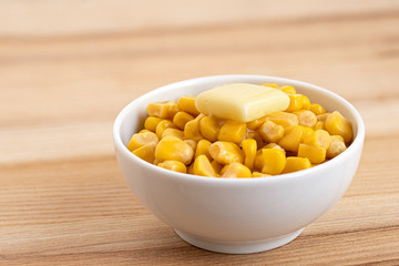 Canned sweet corn with a knob of butter in a white ceramic bowl isolated on light wood. Space for text.