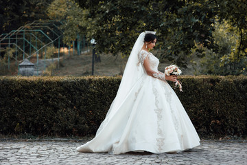 Obraz na płótnie Canvas Bride with a beautiful hair style and crown in hair. The bride in a dress and with a bouquet in her hands for a walk. Tender bride in white wedding dress walking behind nature.