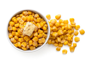 Canned sweet corn with a knob of butter and ground black pepper in a white ceramic bowl next to spilled sweet corn isolated on white. Top view.