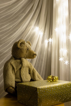 A cute vintage teddy bear with a christmas present wrapped in gold colored paper in front of an illuminated chain of lights