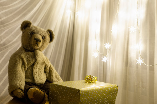 A cute vintage teddy bear with a christmas present wrapped in gold colored paper in front of an illuminated chain of lights