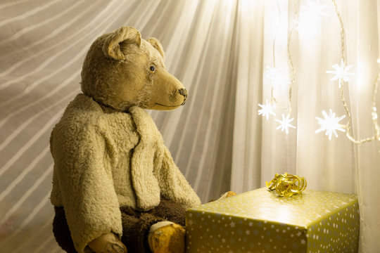A cute vintage teddy bear with a christmas present wrapped in gold colored paper in front of an illuminated chain of lights