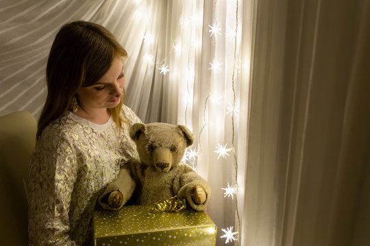 Portrait of a beautiful young white woman holding a teddy bear and a christmas present in her hands in front of an illuminated star shaped christmas chain of lights