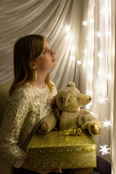 Portrait of a beautiful young white woman holding a teddy bear and a christmas present in her hands in front of an illuminated star shaped christmas chain of lights