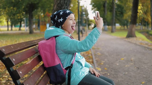 A beautiful teen girl sits on a bench in a park during golden autumn time and takes selfie photos with her mobile phone. Concept of online addiction and social networks.