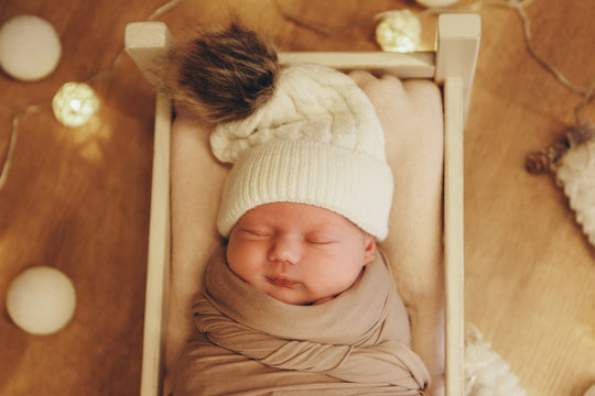 Newborn Baby In A Hat Sleeping In A Crib On The Background Of Christmas Trees . Imitation Of A Baby In The Womb. Portrait Of A Newborn. Healthy Lifestyle Concept, IVF Christmas, New Year Holidays