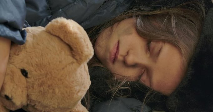 Close-up face of a homeless girl with dirty face sleeping with the teddy bear. Exhausted child wearing hat with earflaps resting on the streets.