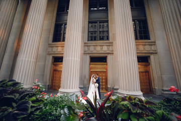 Elegant bride in a white dress and veil. Handsome groom in a blue suit. Couple near large building with columns