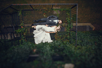 Beautiful bride in a long white dress. Handsome groom in a black suit. Couple lie on a metal