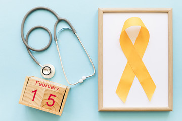 Wooden block calendar showing 15th February, stethoscope and yellow childhood cancer awareness day frame