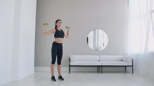 Home exercises for a beautiful body, a young brunette woman lifts dumbbells in her arms up in slow motion