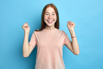 excited teenager with overjoyed expression, rejoices her success, keeps fists raised, , isolated over blue studio background. People, victory and celebration. facial exptionression, positive feeling